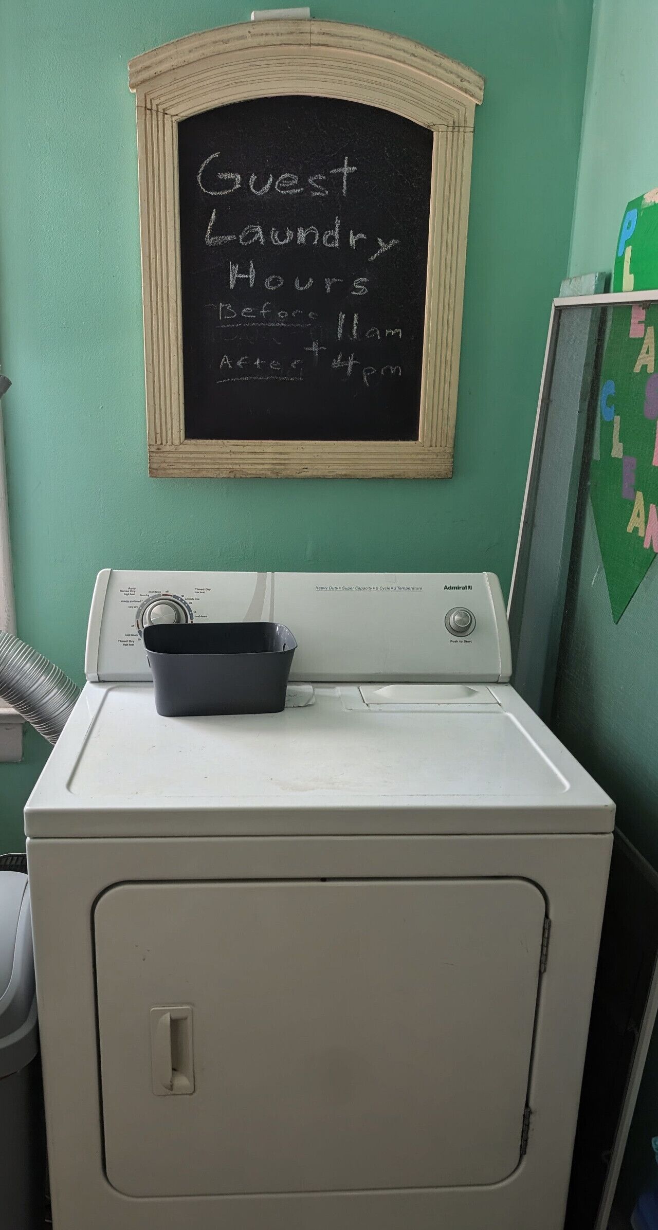 A laundry area with a white washer and dryer, a chalkboard with guest laundry hours, and a green wall.