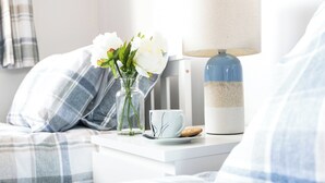 A neatly made bed with a plaid blanket, a bedside table with a lamp, a cup, and a vase with flowers. - 3 bedroom accommodation in Sheringham (Sheringham)