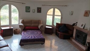 A living room with a beige sofa, a patterned ottoman, a brown leather sofa, and a fireplace. There are two windows with arched tops and a view of greenery outside. - "La Favorita: Mountain Serenity with Ocean Views". (Calpe)