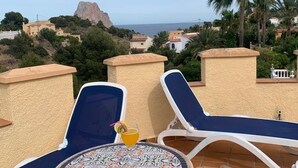 A terrace with two lounge chairs and a table, overlooking a coastal town with a prominent rock formation and palm trees. - "La Favorita: Mountain Serenity with Ocean Views". (Calpe)