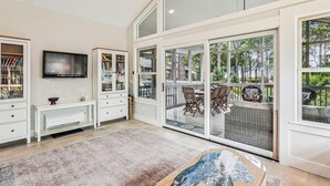 A spacious living room with a large TV, built-in bookshelves, a dining area with outdoor views, and a glass door leading to a balcony.