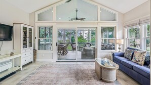 A spacious living room with a high ceiling, large windows, a ceiling fan, a television, a sofa, and a coffee table.