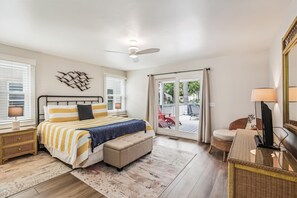 Un dormitorio con cama, mesitas de noche, ventilador de techo, ventana grande y puerta corrediza de vidrio que da a un espacio exterior.