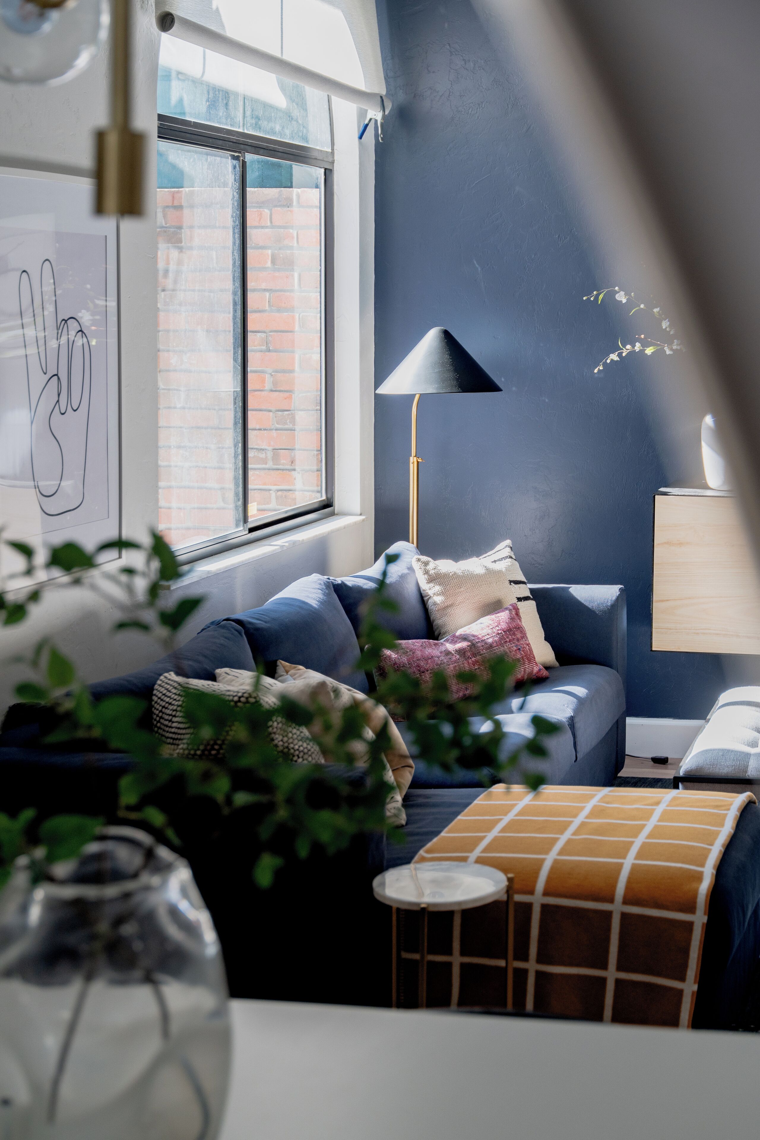 A modern living room with a blue sofa, a small round table, and a lamp. A window with a view of a brick wall is visible.