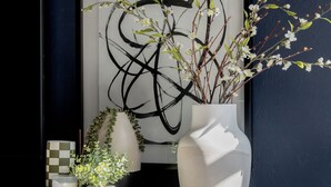 Een houten kast met een witte vaas met takken en kleine witte bloemen, een lijst met een abstract schilderij, en een geruit object op een dienblad.
