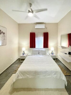 A hotel room with a large bed, two bedside tables with lamps, a ceiling fan, a window with red curtains, and a mounted air conditioner. - Dawn Beach Club 2 Bed…2Bath Villa, With Beach Access & Resort Bar (Upper Prince's Quarter)