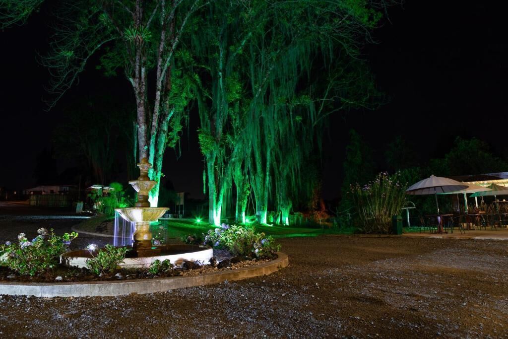 Un espacio al aire libre iluminado por la noche con una fuente, árboles y una zona de estar bajo un toldo.