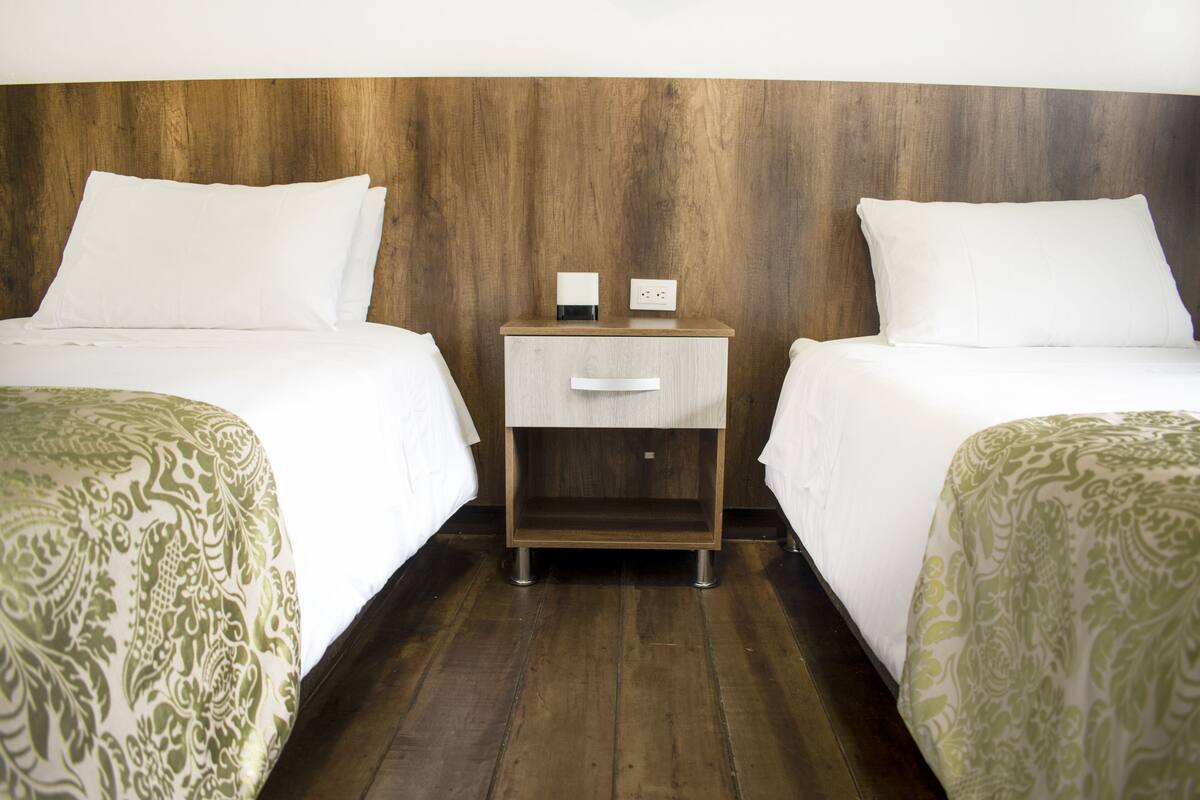 A hotel room with two beds, a wooden headboard, a nightstand with a lamp, and a drawer.