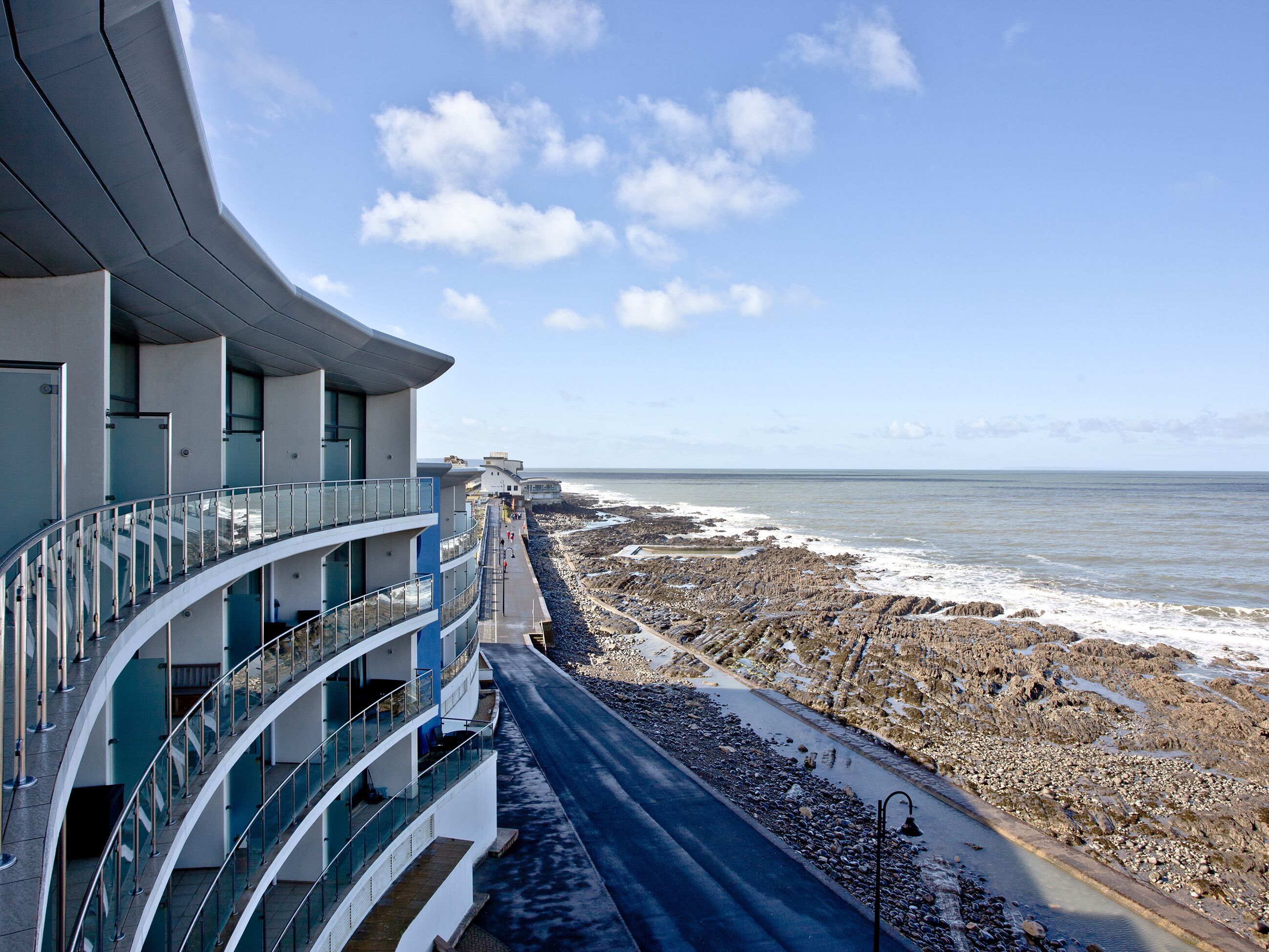 Um edifício moderno com varanda curva com vista para uma praia rochosa e o mar.