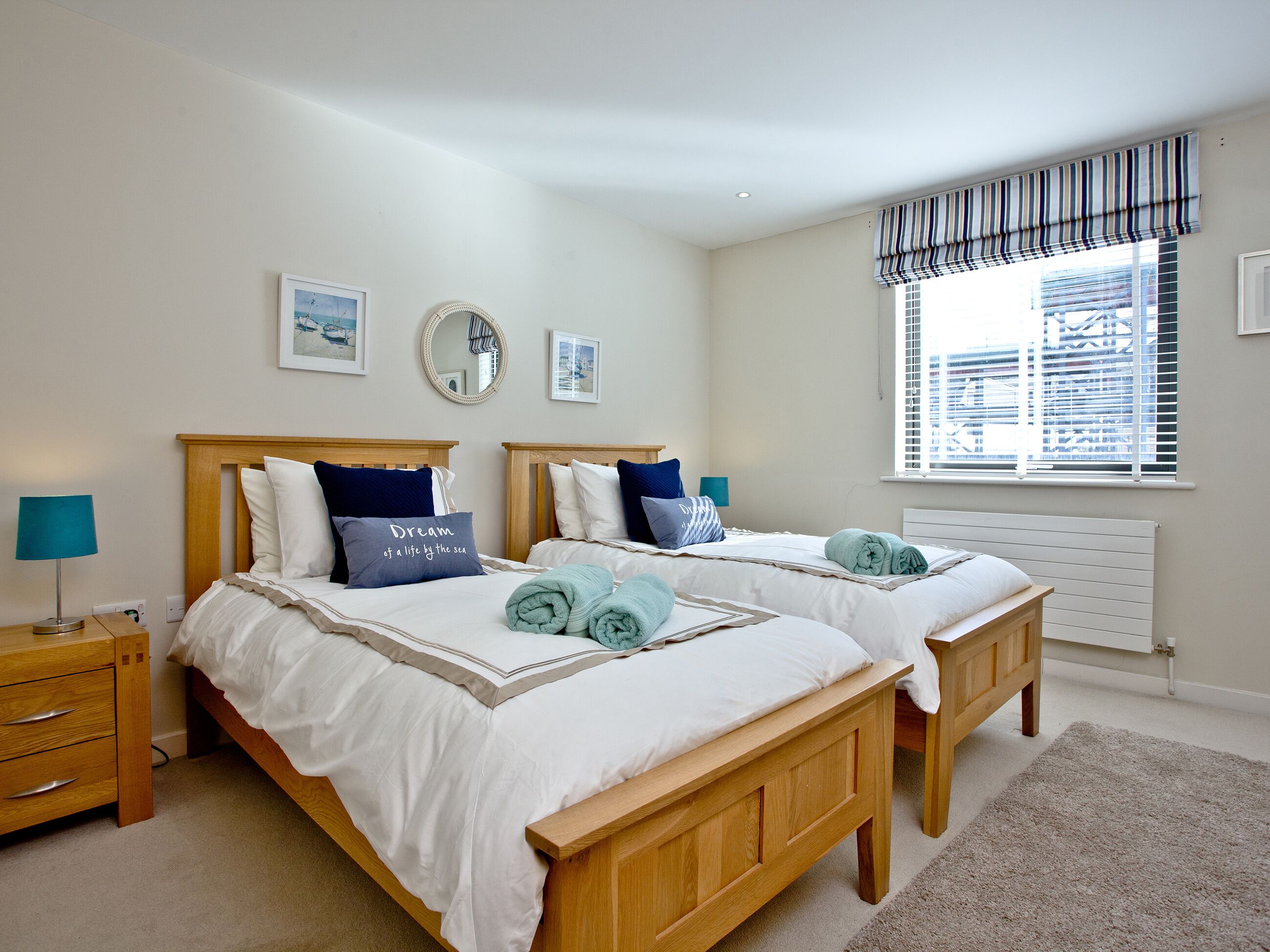 A double bedroom with two beds, bedside tables, a window with blinds, and a view of the city.