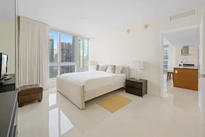 A modern hotel room with a large bed, a bedside table, a lamp, a small ottoman, and a view of the city through large windows.