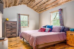 A bedroom with a wooden ceiling, a bed with pink bedding, two windows with patterned curtains, and a wooden dresser.
