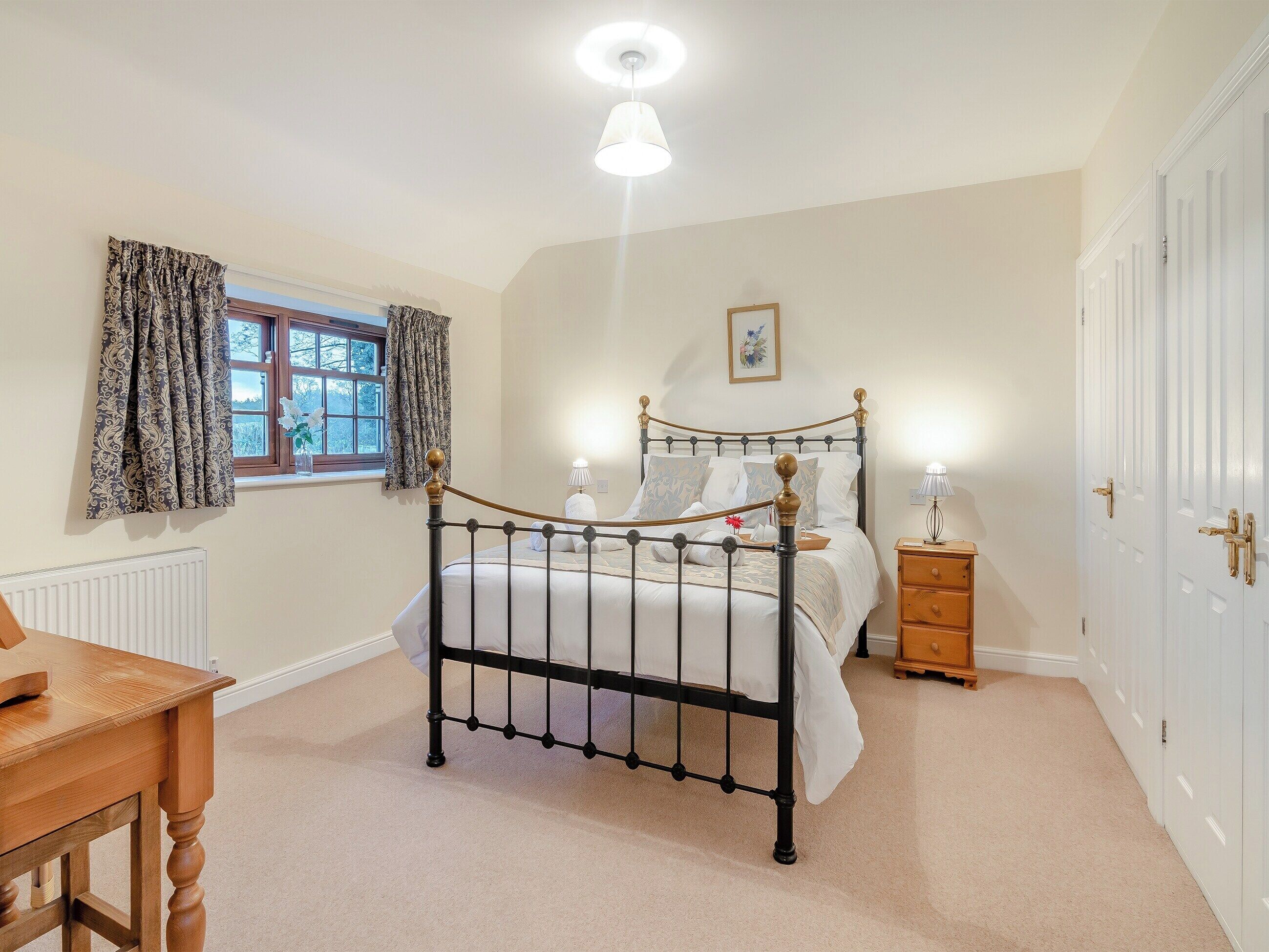 A bedroom with a four-poster bed, a bedside table, a nightstand, a radiator, a window with curtains, and a door.