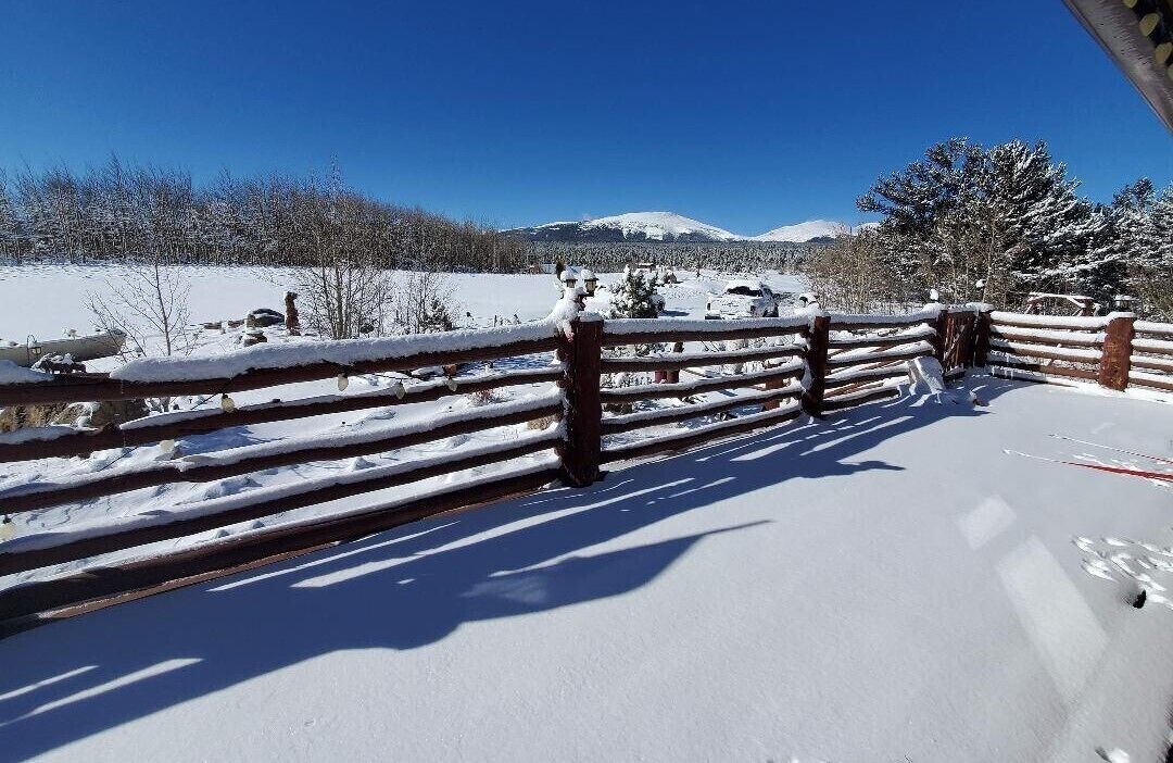 A snow-covered outdoor area with a wooden fence, a clear blue sky, and a mountain in the background.