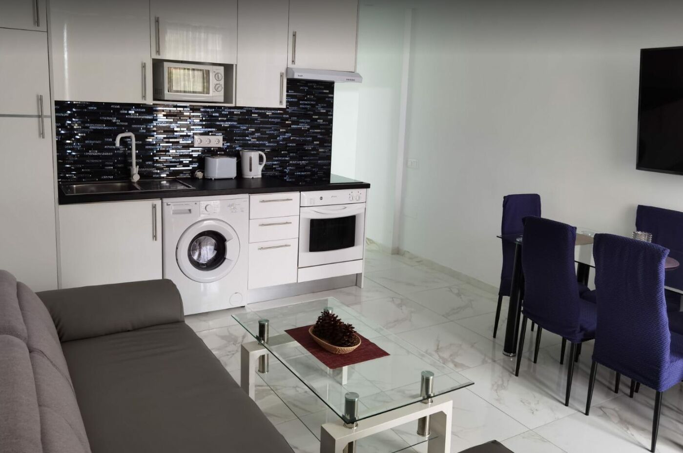 A modern kitchen with a washing machine, oven, and microwave. There is a dining area with purple chairs and a glass table.