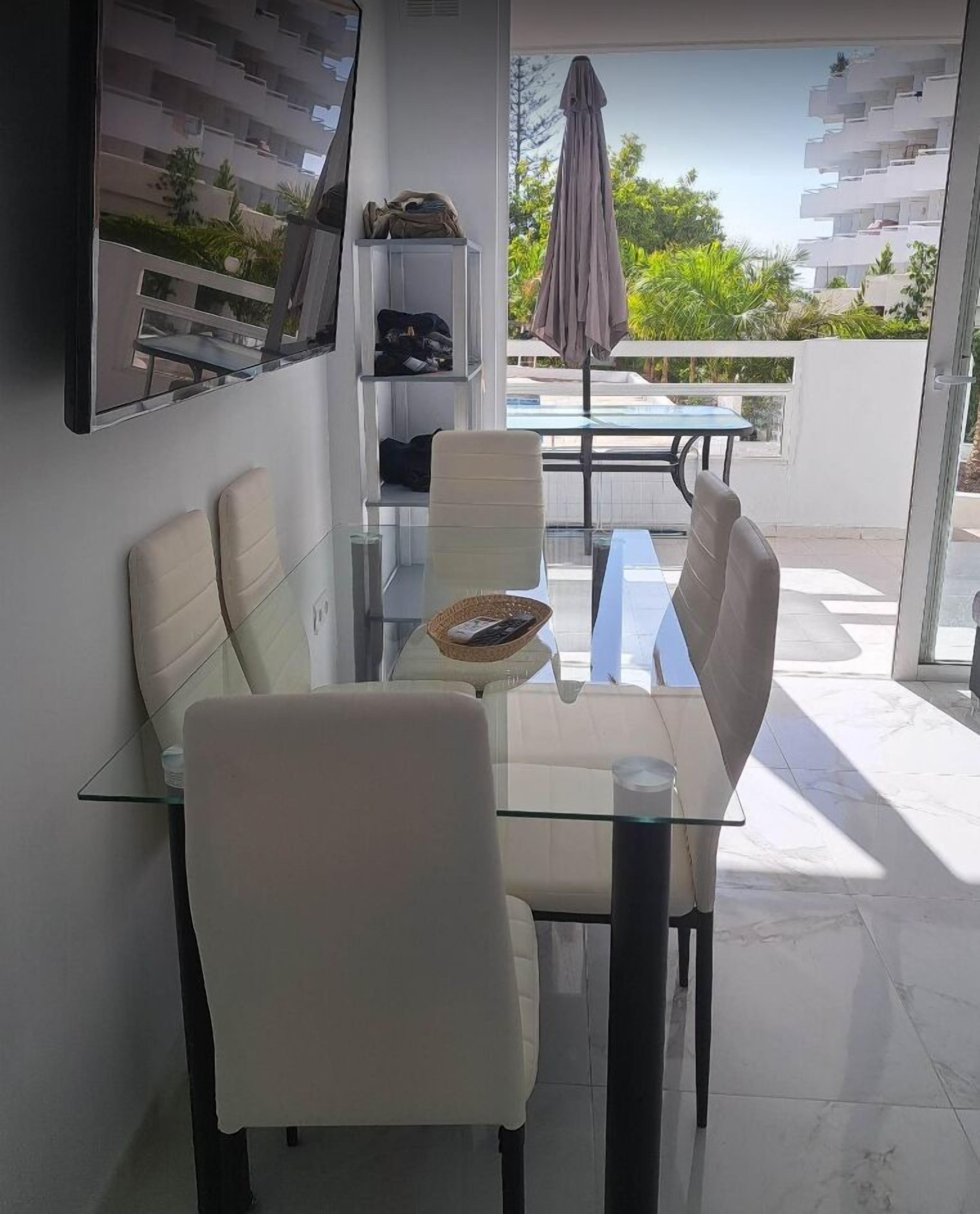 Un espace repas moderne avec une table et des chaises en verre, une petite table d’appoint avec un panier, et une vue sur un balcon équipé d’une table et de chaises.