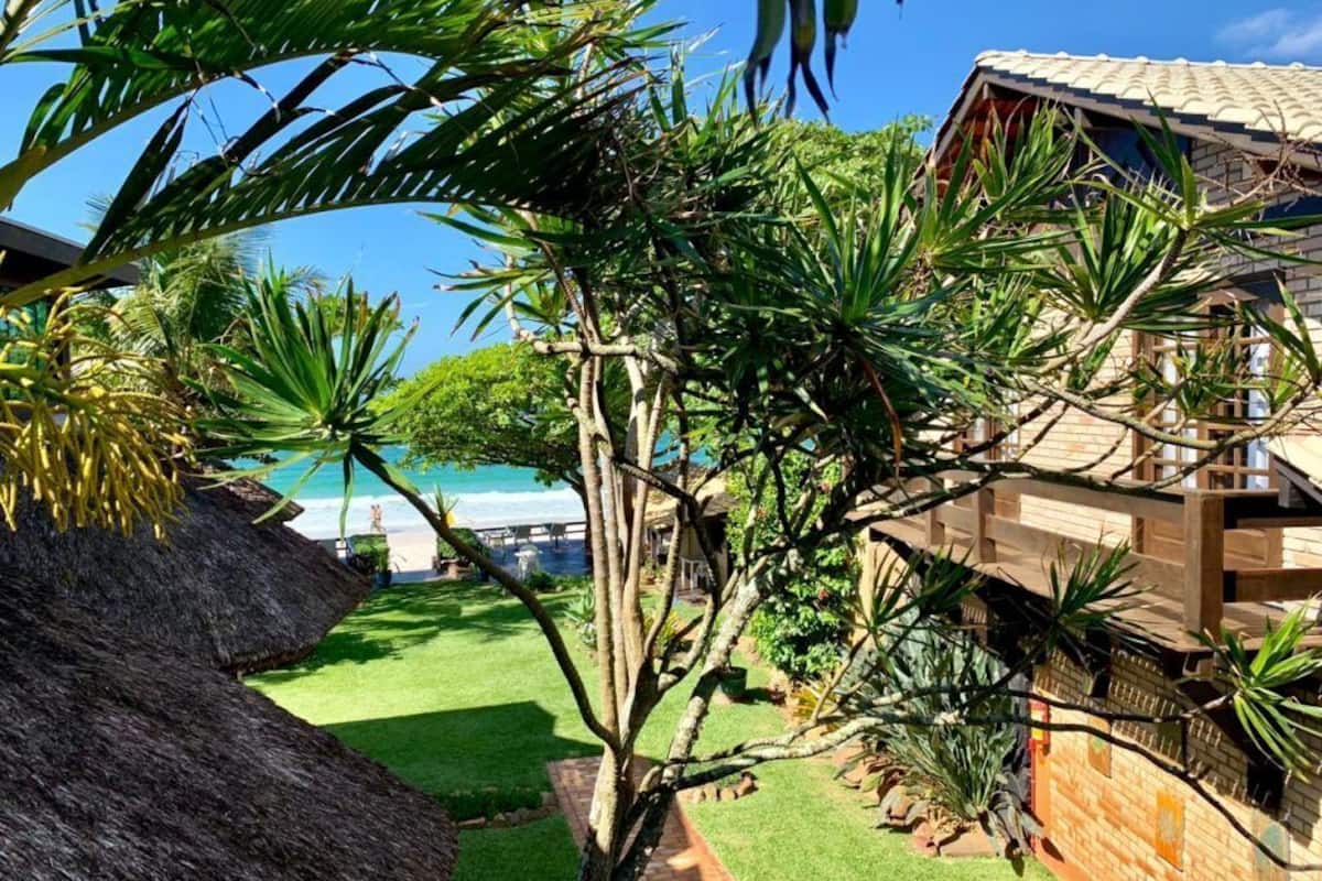A tropical garden with a thatched roof structure, a brick building, and a view of the ocean.