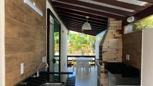 A modern kitchen with a central island, a dining area with a table and chairs, and a view of a lush garden. - Pousada Solar dos Girassóis (Bombinhas)