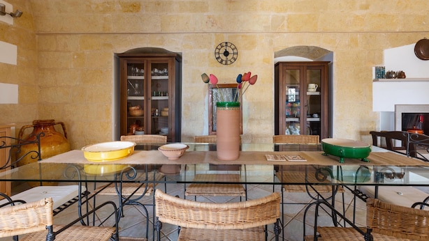 A dining area with a glass table, wicker chairs, a large vase with flowers, and stone walls. - 2920 Villa Oasis by Perle di Puglia (Mottola)