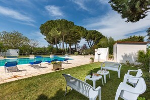 A pool area with blue lounge chairs, a white table with a bottle and glasses, and a small white building.