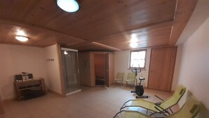 Ein Zimmer mit Holzboden, einer Glastür zu einem weiteren Raum, einem Laufband und mehreren Stühlen.