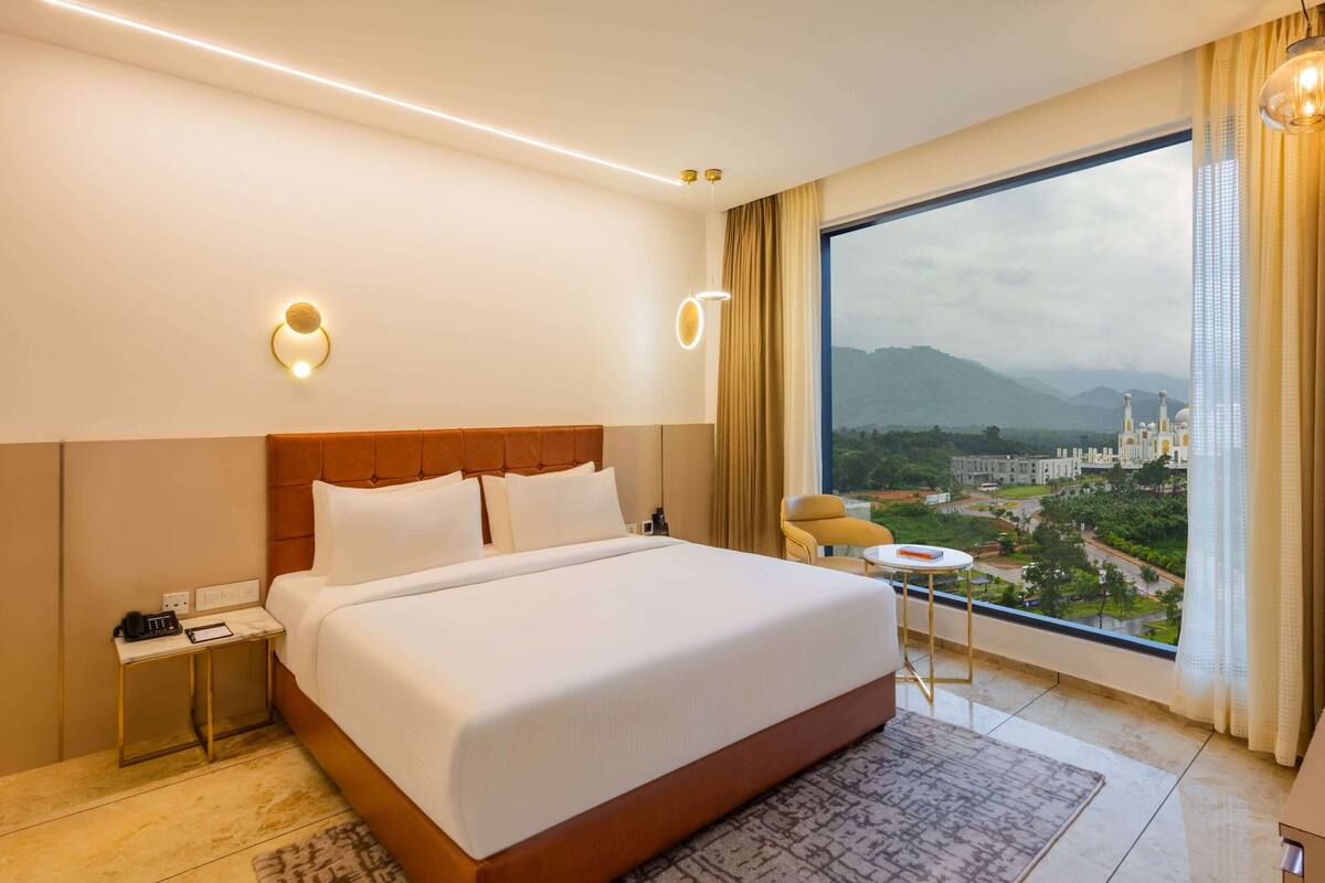 Habitación de hotel con una cama grande, mesita de noche y vista a un paisaje urbano.