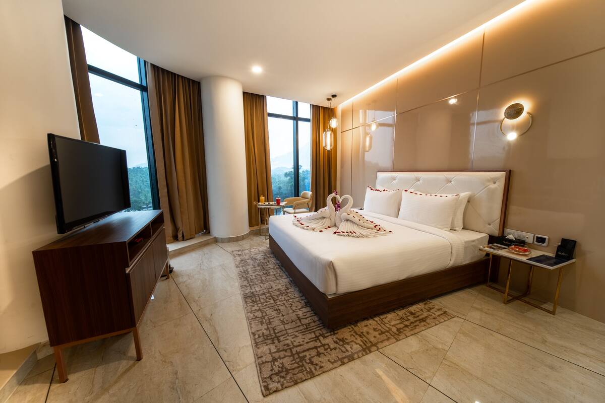 Una habitación de hotel moderna con una cama grande, un televisor sobre un mueble de madera, una mesita con una silla, y vistas al exterior a través de amplios ventanales.