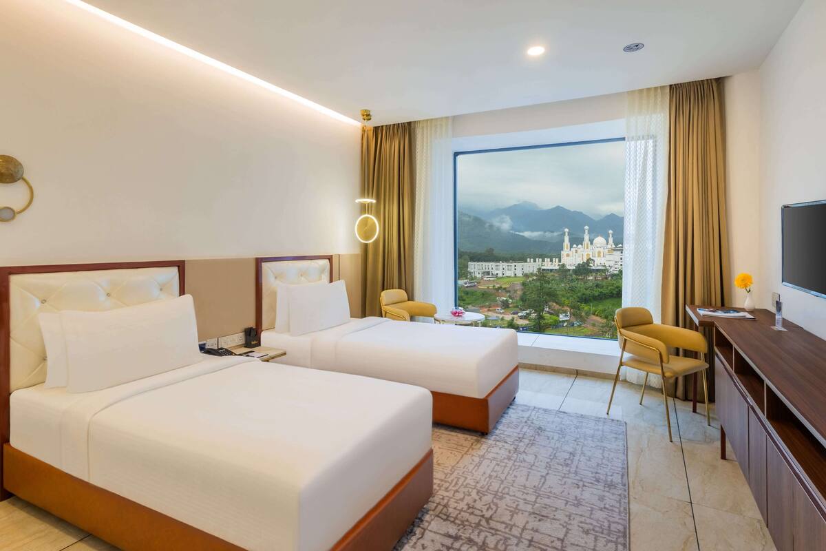 Habitación de hotel con dos camas, un escritorio, una silla, un televisor y un amplio ventanal con vistas a un edificio y montañas.