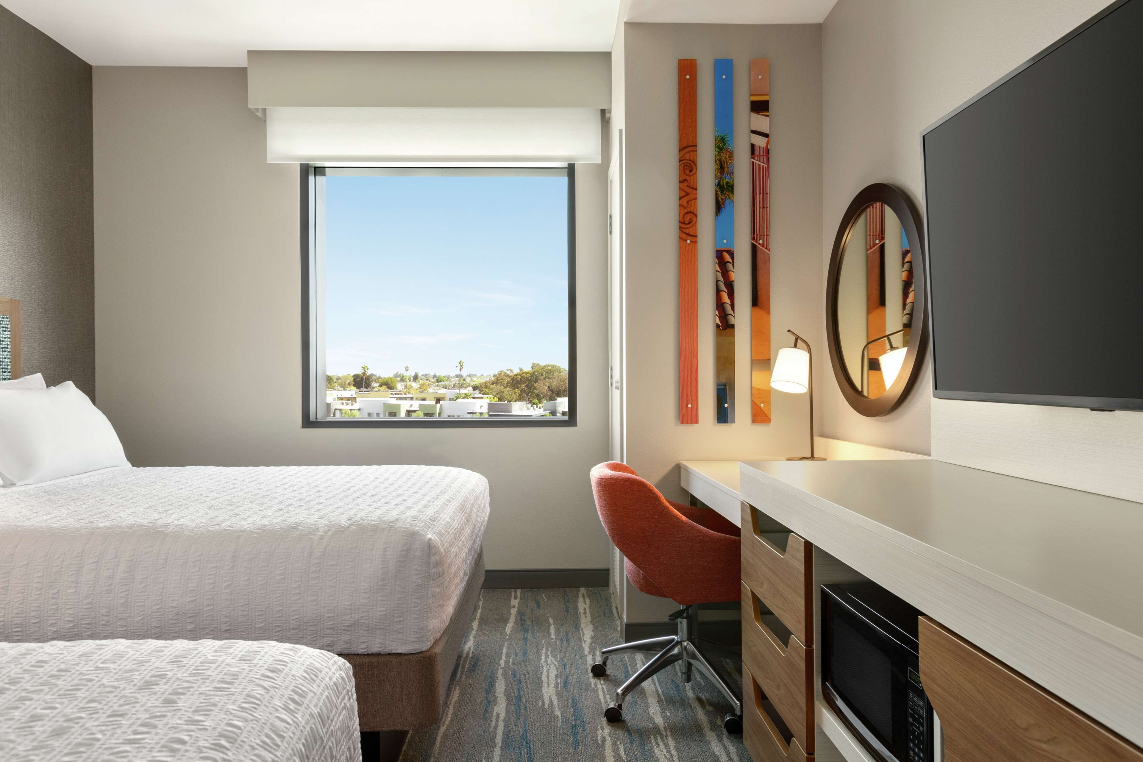 Habitación de hotel con dos camas, un escritorio, una silla, un televisor de pantalla plana, un ventanal grande y vistas a la ciudad.