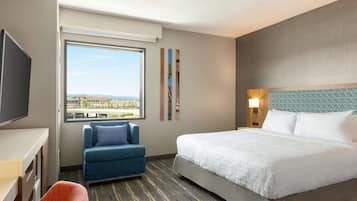 Ein Hotelzimmer mit einem großen Bett, einem Schreibtisch, einem blauen Sessel und Blick auf eine Stadtlandschaft durch ein großes Fenster.