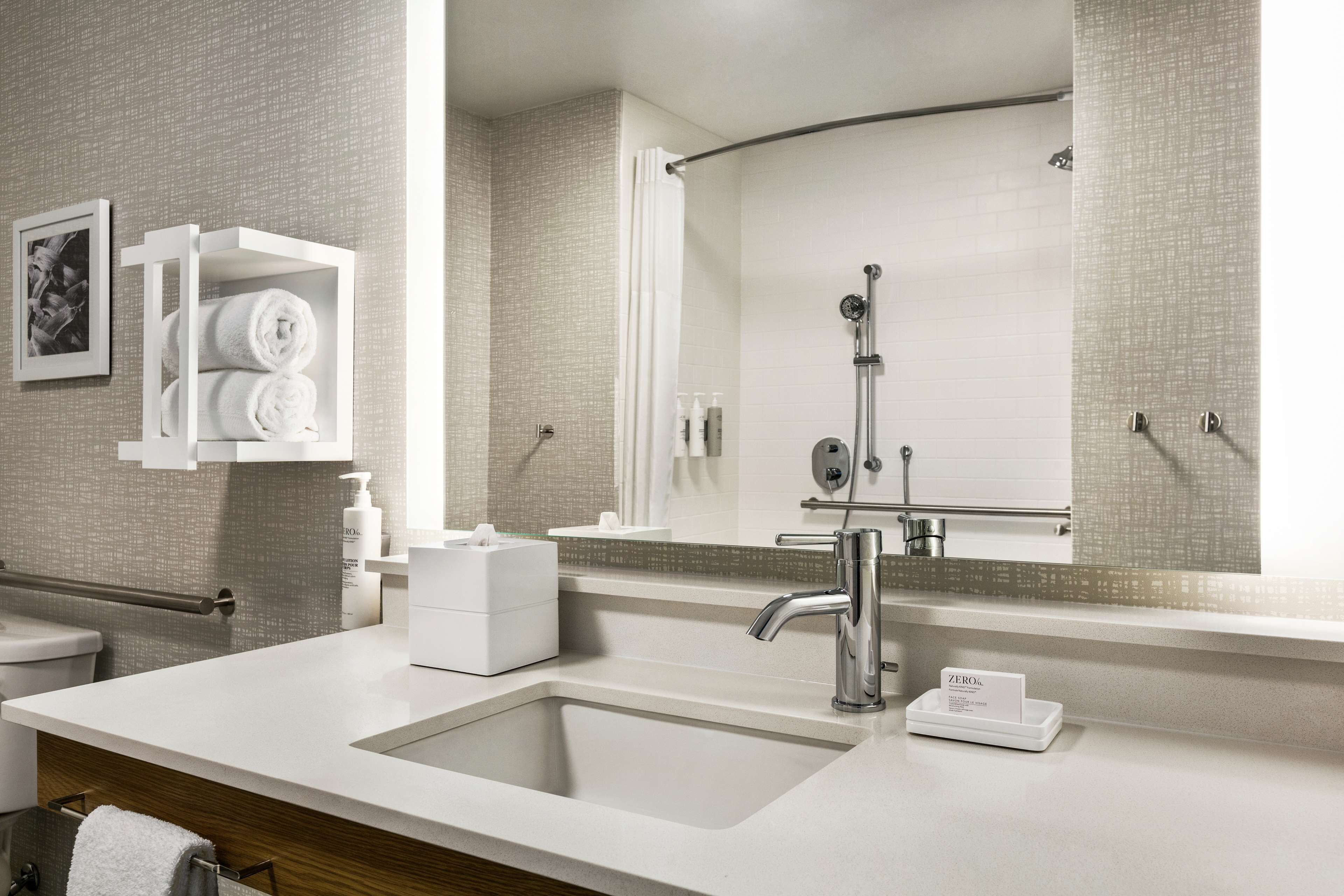 Une salle de bain moderne avec un grand lavabo, un miroir et une douche. On y trouve un porte-serviettes avec des serviettes et une petite boîte blanche posée sur le comptoir.