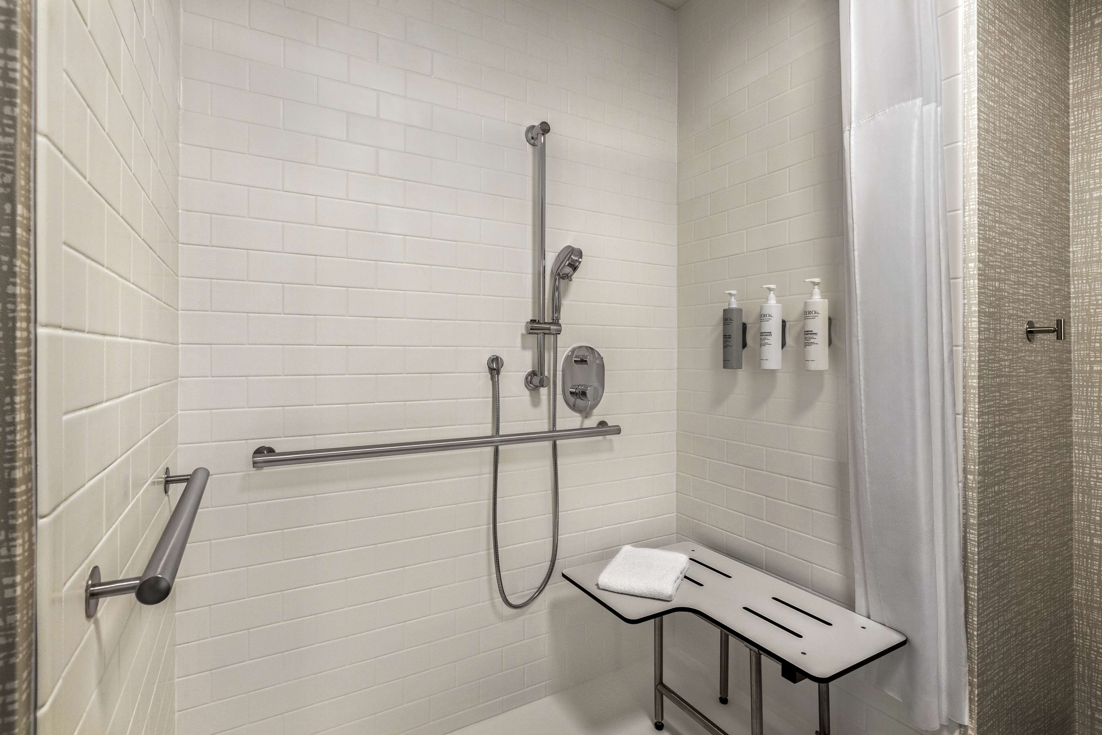 A modern bathroom with white tiled walls, a shower with a handheld showerhead, a grab bar, and a bench with a towel.