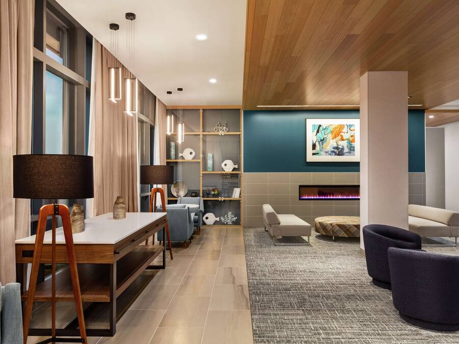 Een moderne hotel lobby met een houten plafond, een groot raam met gordijnen, een zithoek met banken en stoelen, een tafel met een lamp en een open haard.
