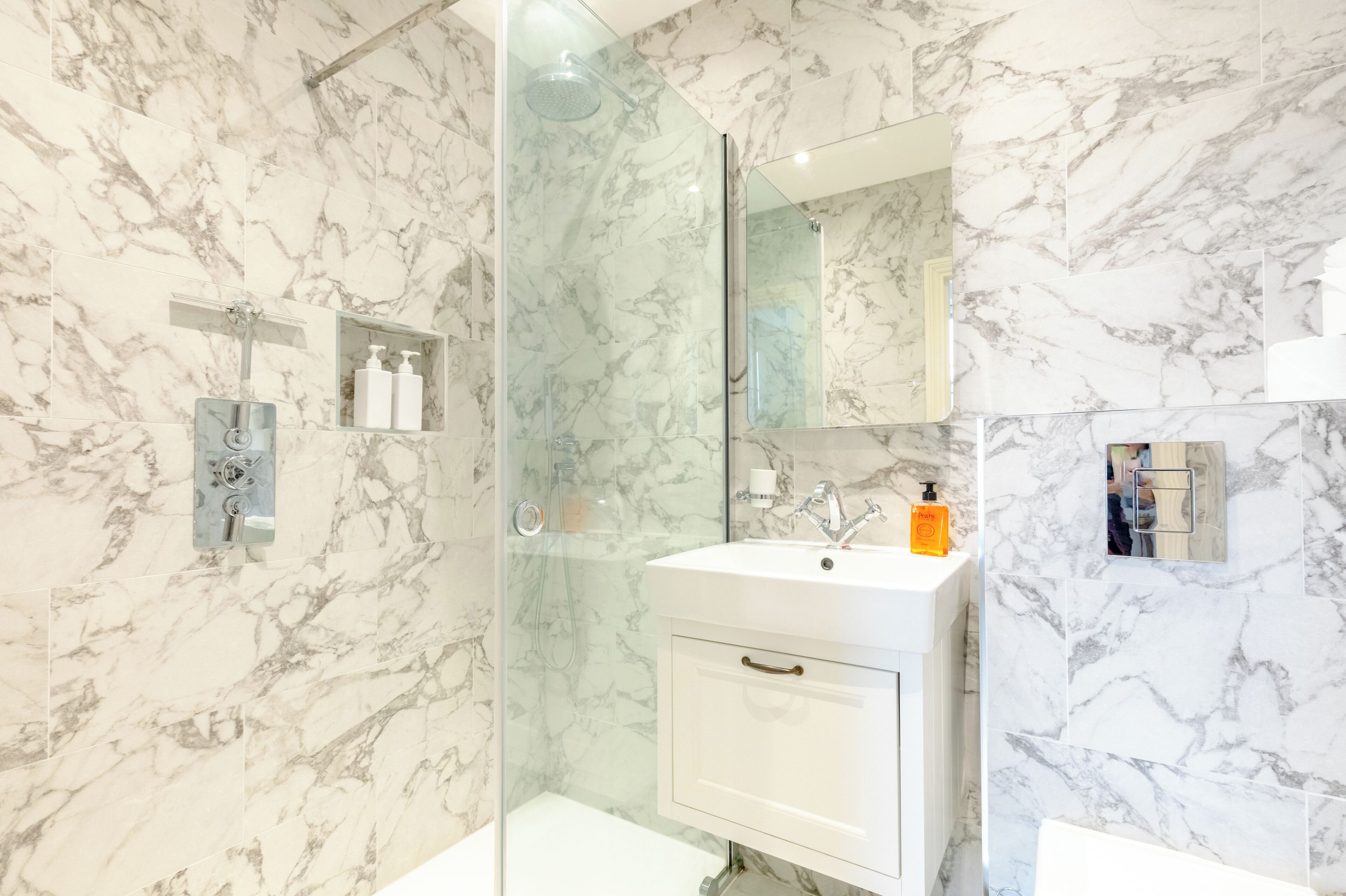 A bathroom with marble walls, a glass-enclosed shower, a white sink with a mirror above it, and a small shelf with toiletries.