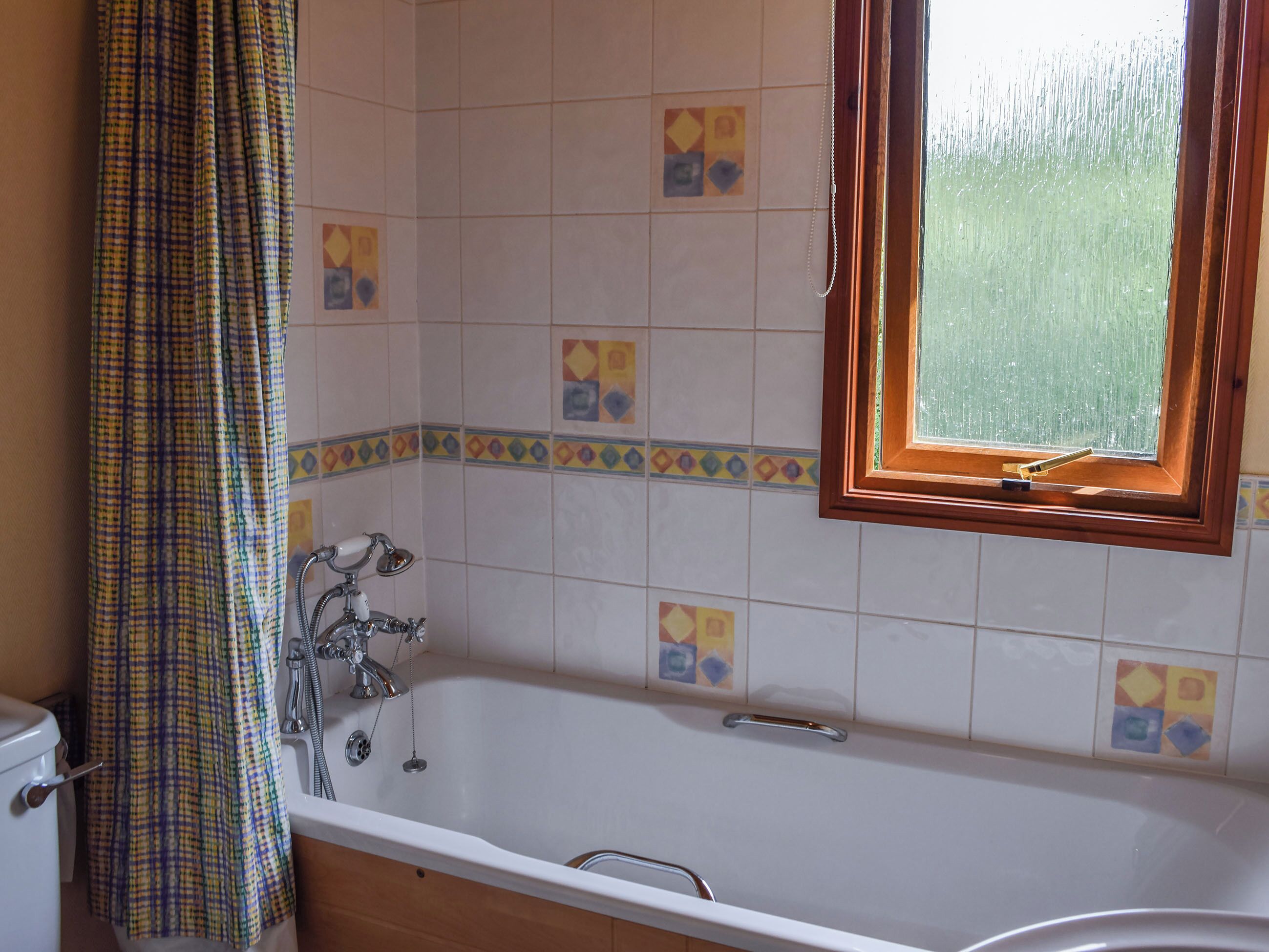 Et badeværelse med badekar, bruser og toilet. Der er et vindue med træramme og gardin.