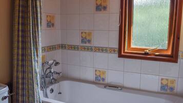 Et badeværelse med badekar, bruser og toilet. Der er et vindue med træramme og gardin.