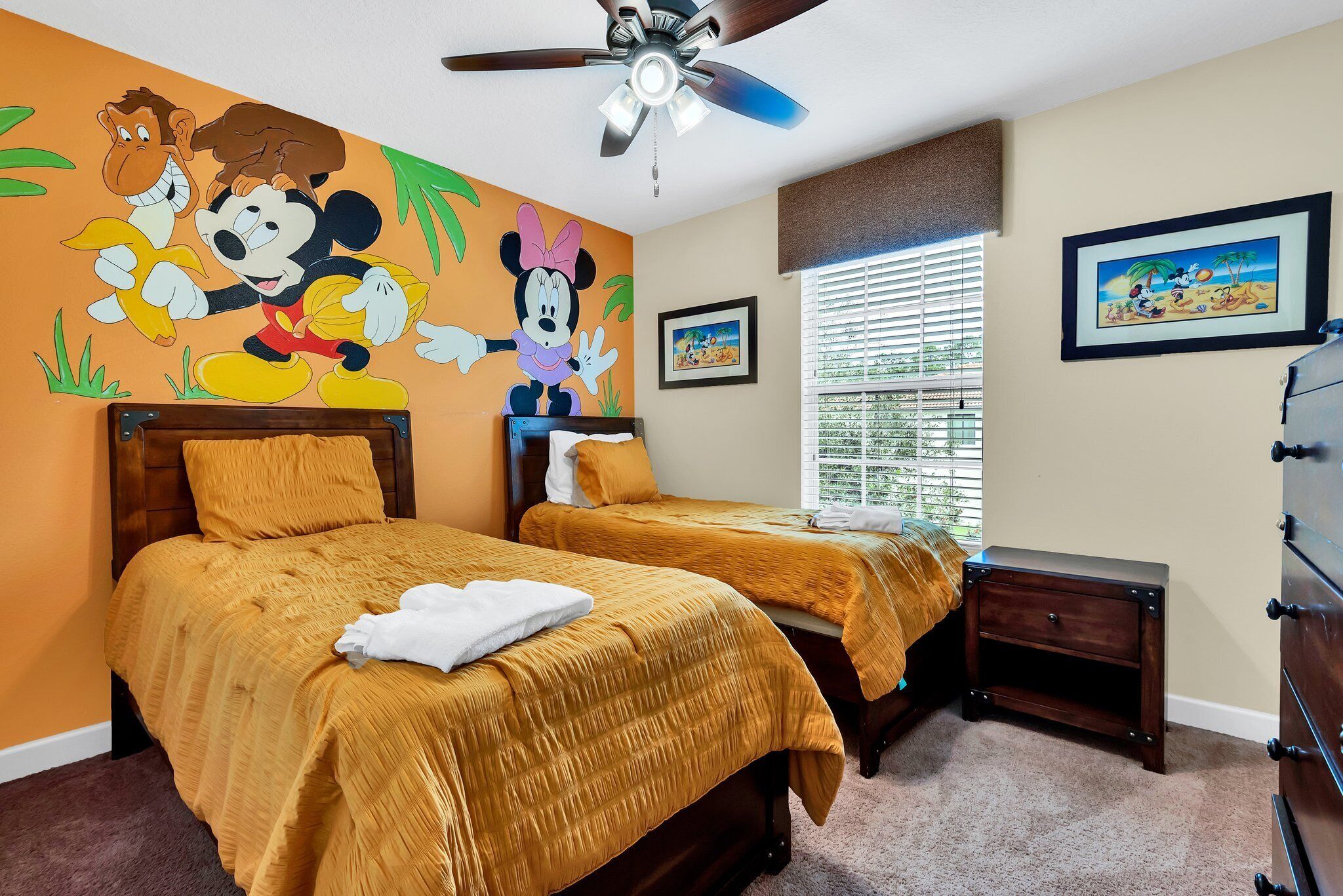 A hotel room with two beds, a ceiling fan, a window with blinds, and a wall with Disney-themed artwork.