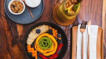 Uma mesa de madeira com um prato de frutas, um recipiente de granola, um copo de suco e um prato com uma fatia de pão e tomate.