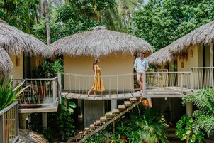 Una pareja caminando sobre un puente de madera entre dos cabañas con techo de paja en un exuberante jardín.