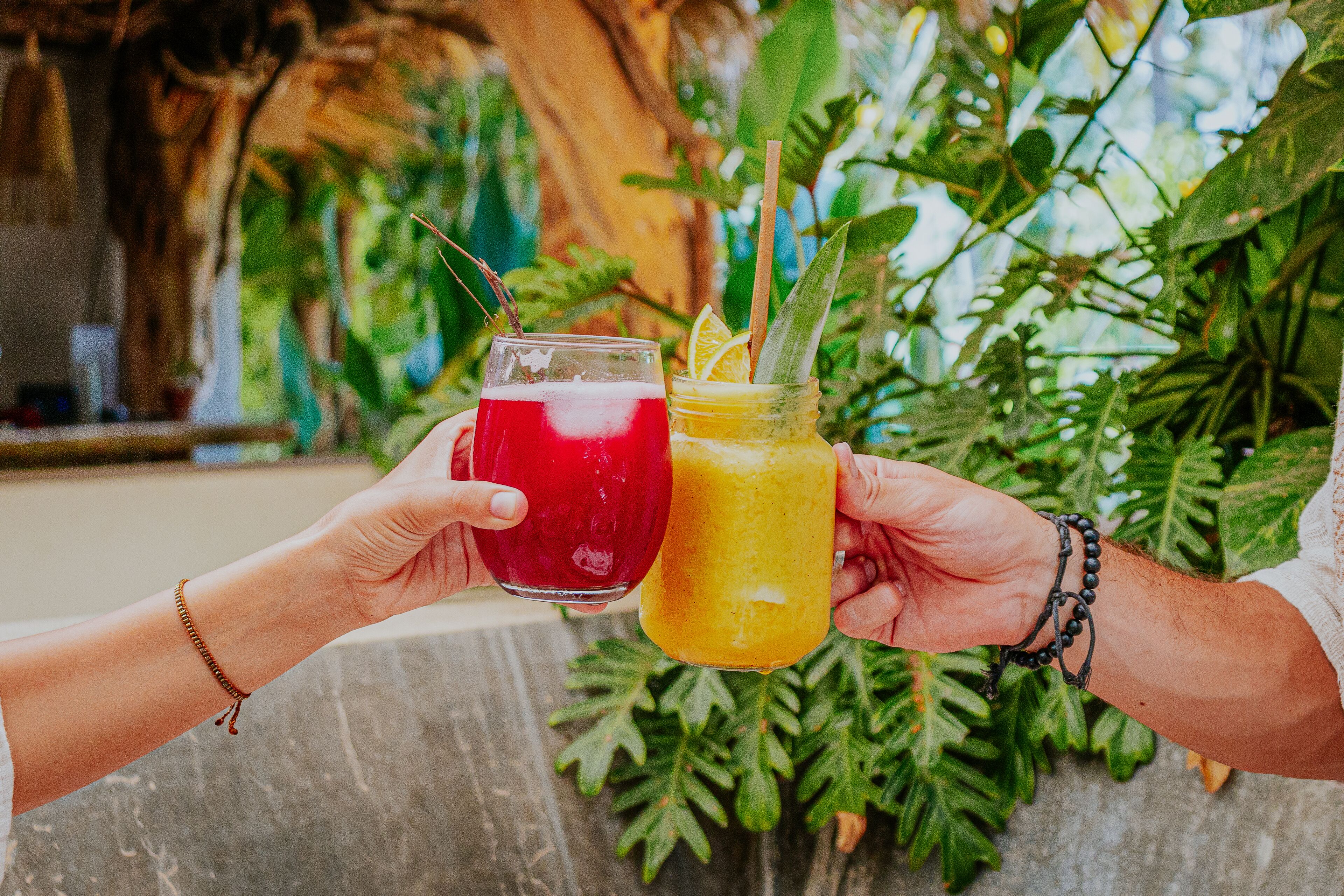 Two people holding drinks with straws, one red and one yellow, in a tropical setting with lush greenery.