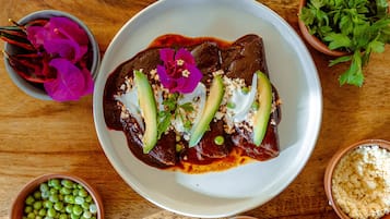 Plato de enchiladas cubiertas de aguacate, queso y salsa, rodeado de tazones con ingredientes como frijoles, cebollas y cilantro.