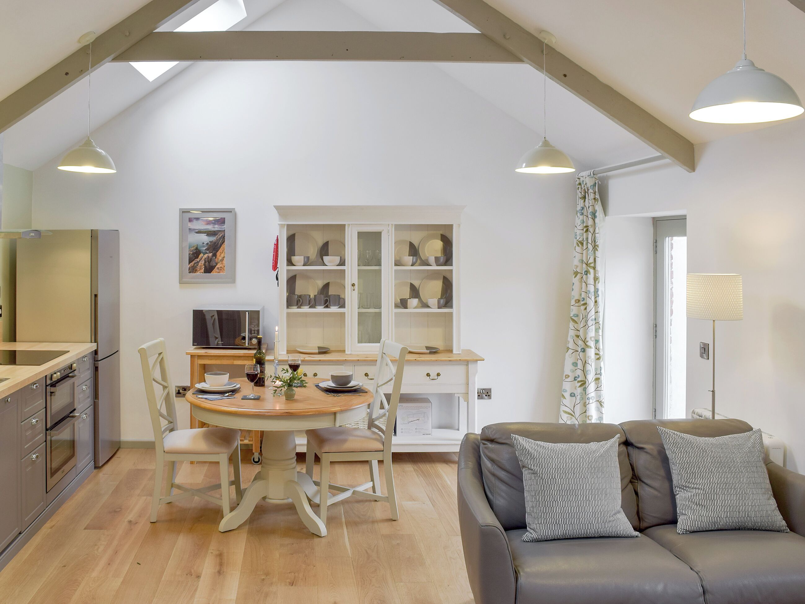 A modern kitchen with a dining table, chairs, and a sofa in a bright room with wooden beams.