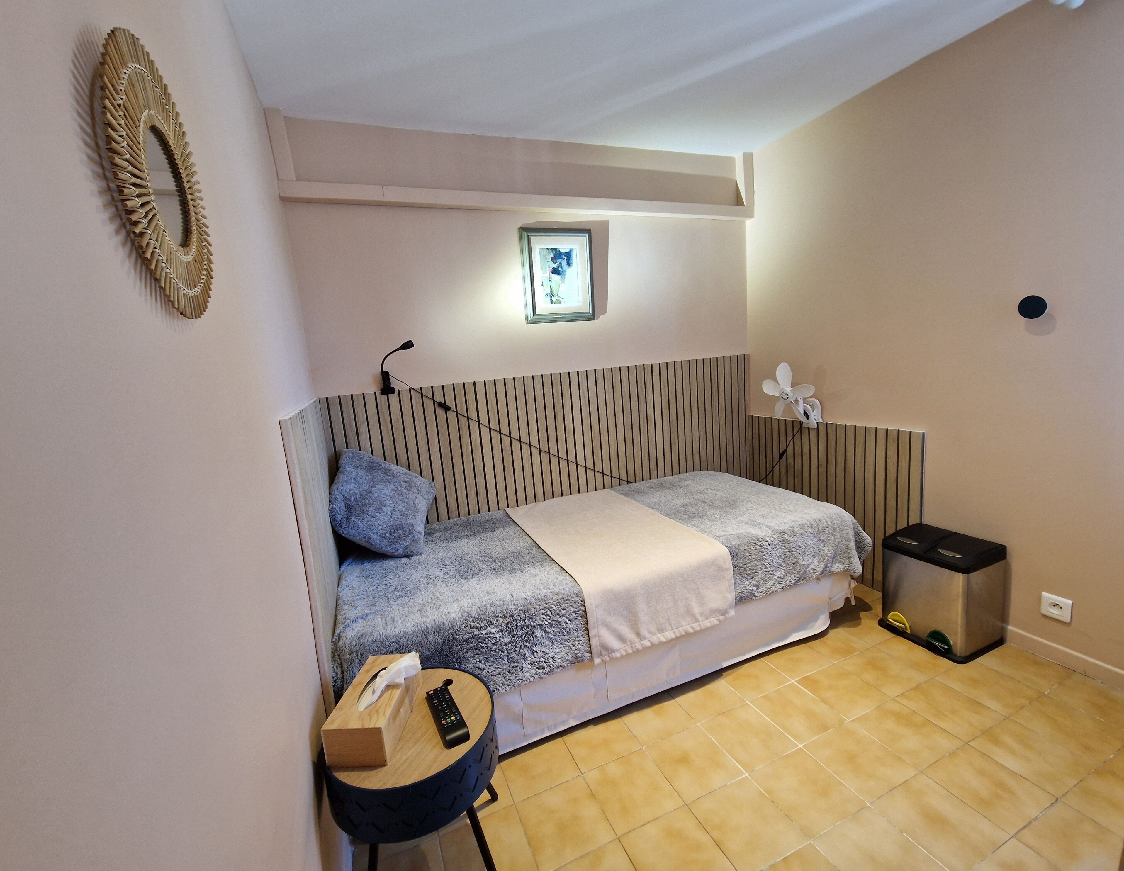 Una camera spaziosa con un letto, un comodino, un cestino, una ventola a parete e un quadro incorniciato.