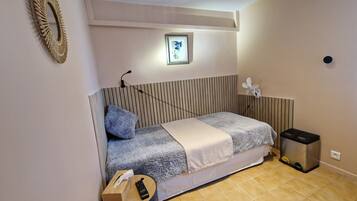 Una camera spaziosa con un letto, un comodino, un cestino, una ventola a parete e un quadro incorniciato.