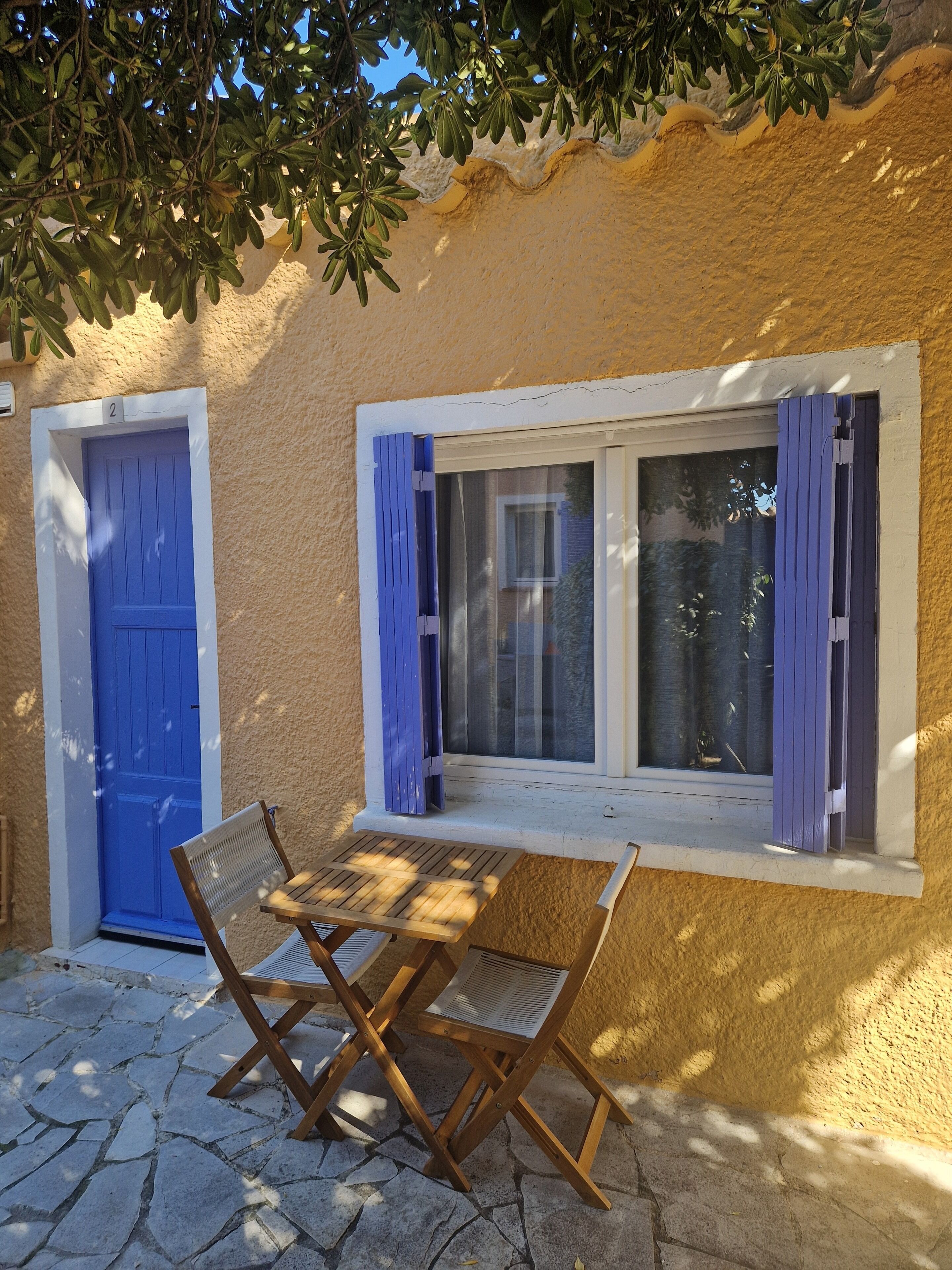 A room with a stone floor, a blue door, a window with purple curtains, and a wooden table with chairs.