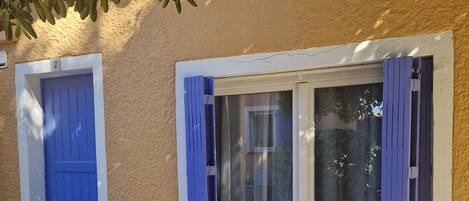 A room with a stone floor, a blue door, a window with purple curtains, and a wooden table with chairs.