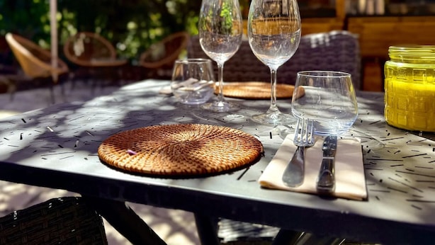 A table set outdoors with wine glasses, a woven placemat, cutlery, and a yellow container, likely containing a drink. - le Cosy (Fréjus)