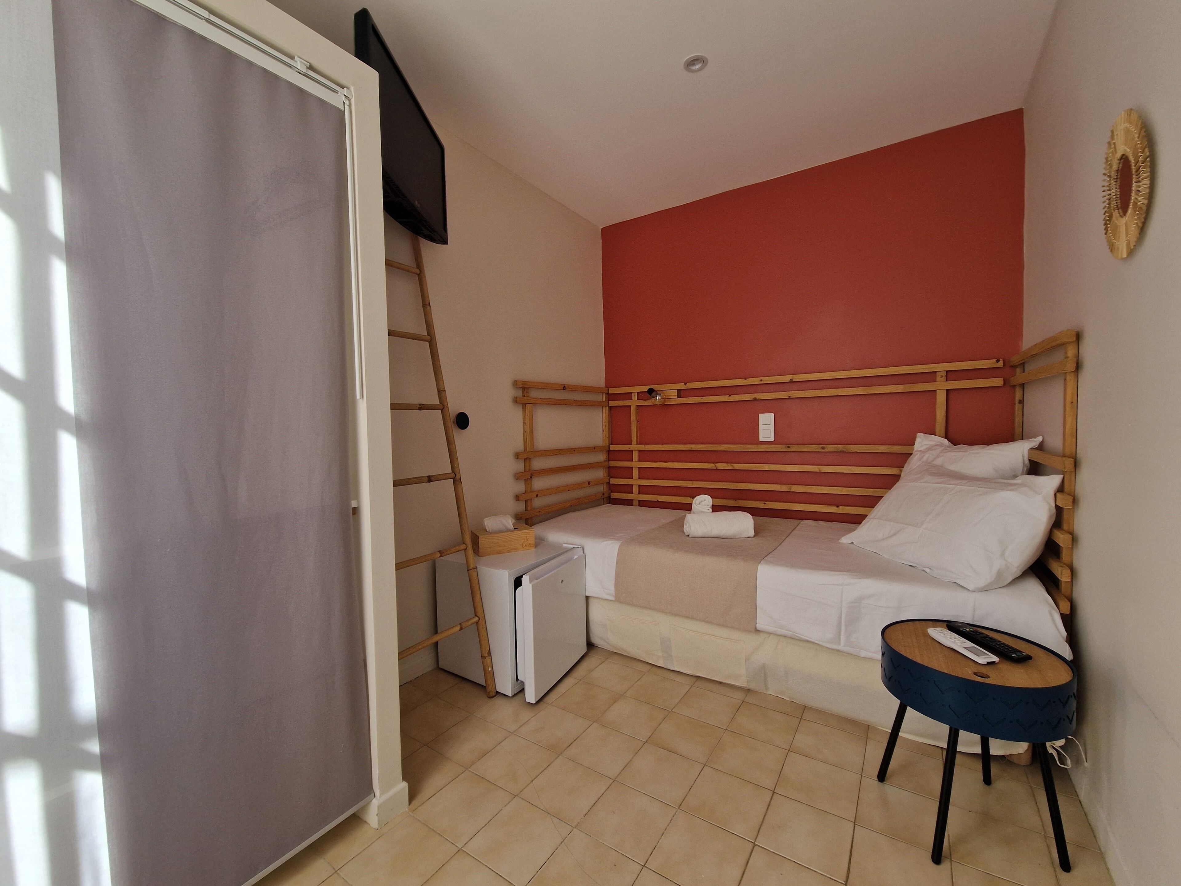 A small room with a bed, a small table, a ladder, and a television mounted on the wall.