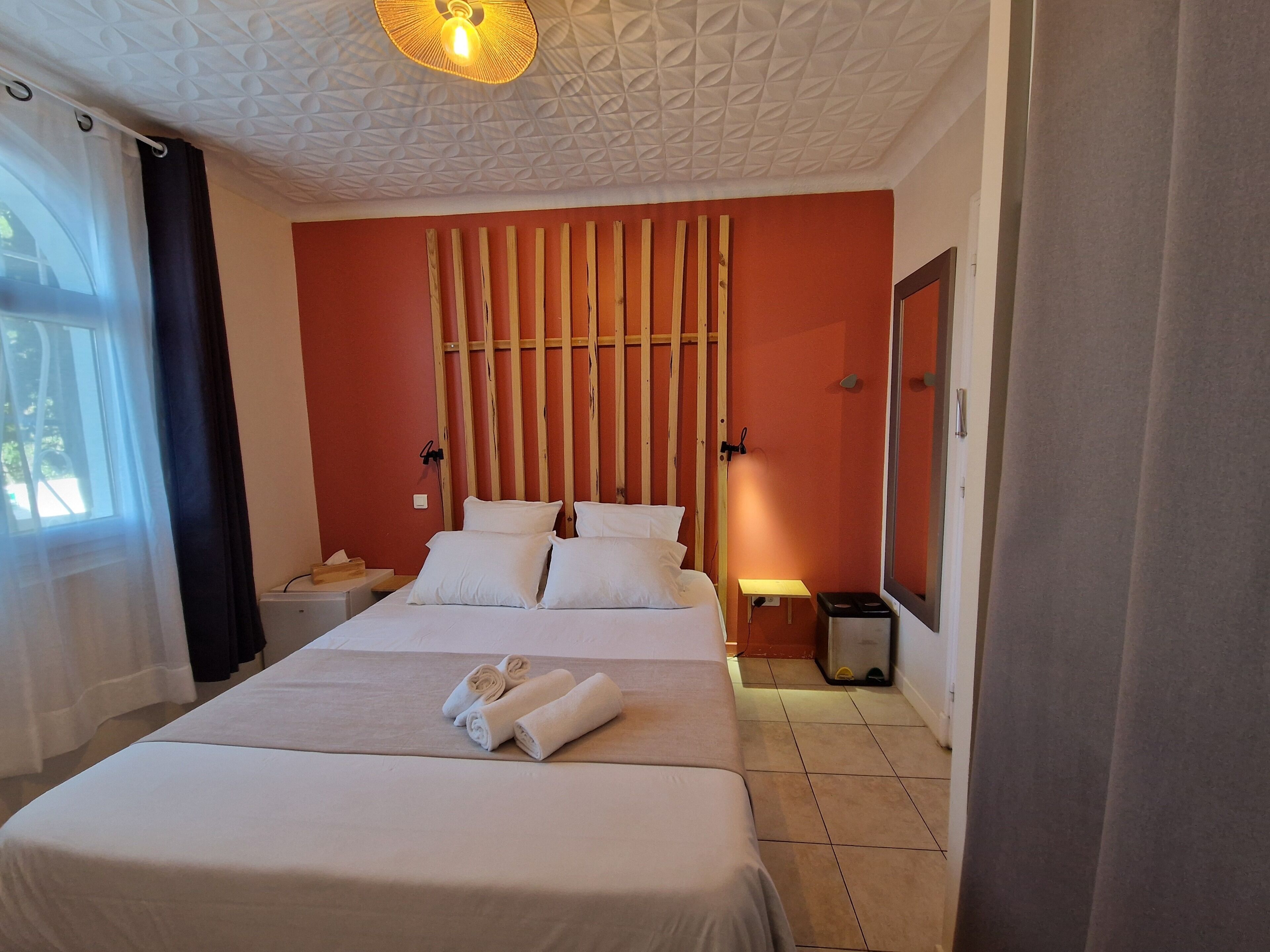 Une chambre d’hôtel comprenant un lit, une table de chevet, une commode, un miroir et une fenêtre avec des rideaux.