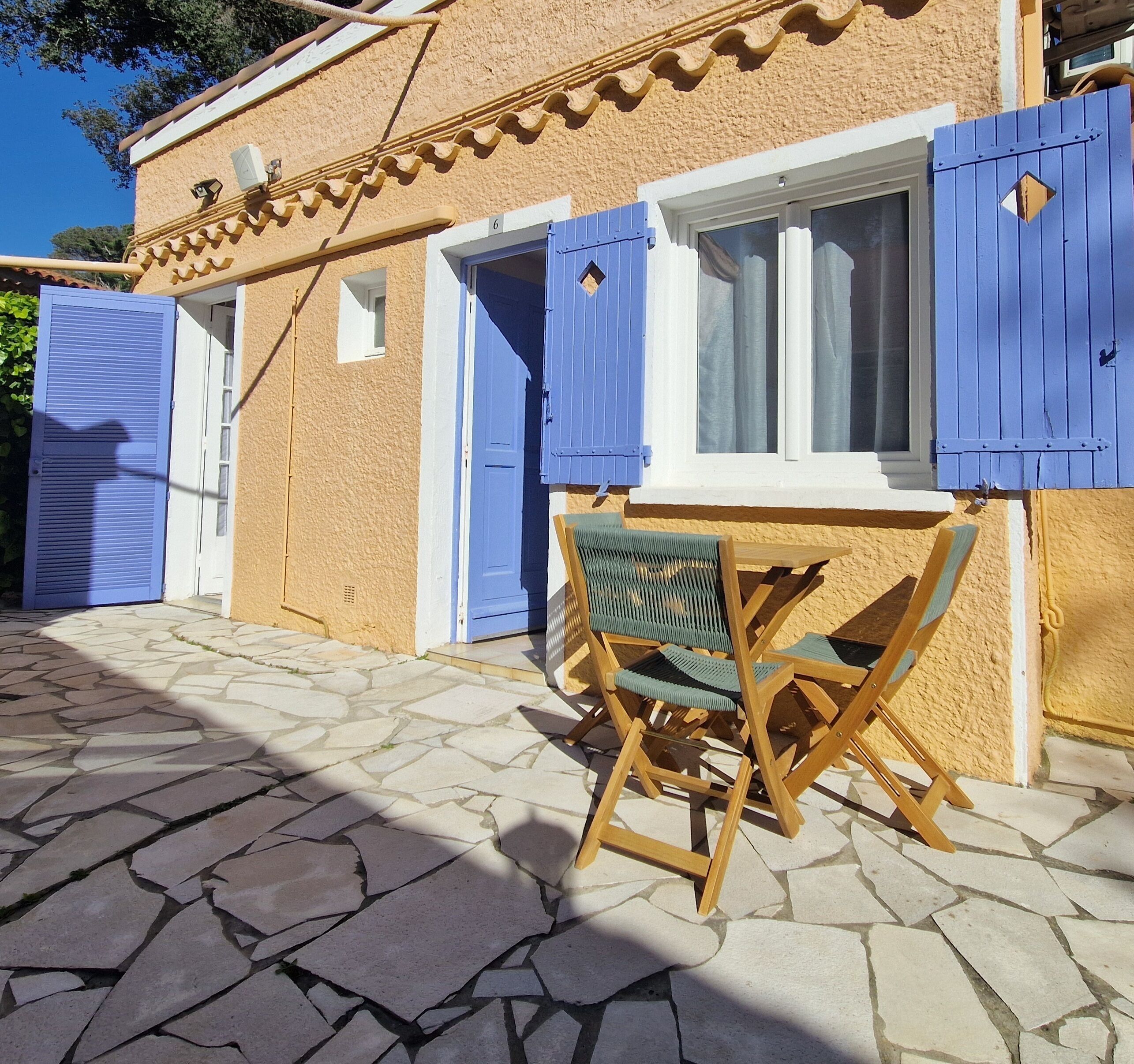 A stone-paved courtyard with a wooden table and chairs, a window with blue shutters, and a door with a blue shutter.